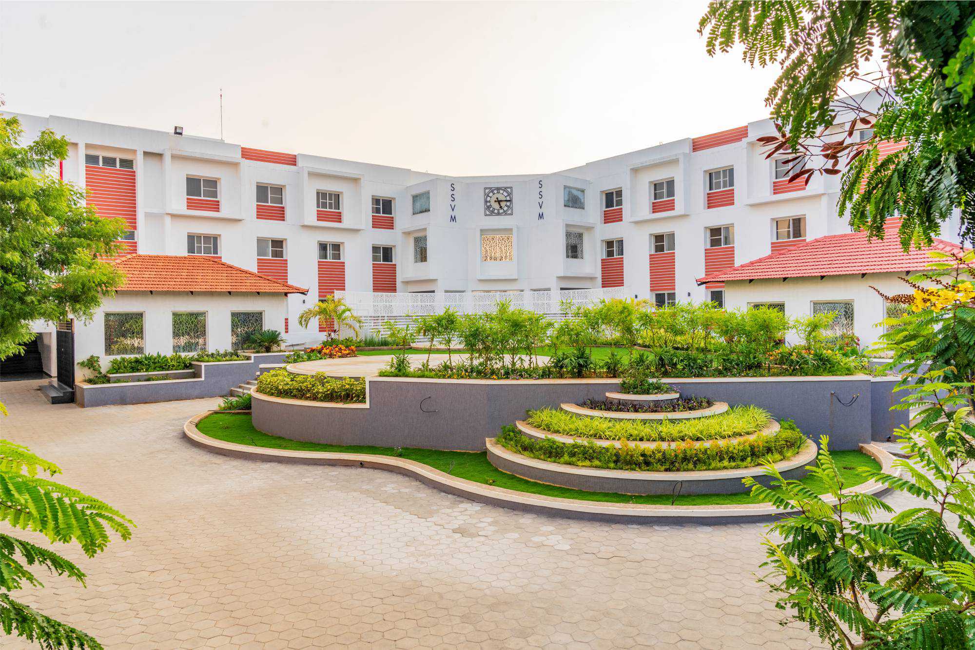 A white, multi-story building with red accents, featuring a central courtyard with tiered gardens and well-maintained landscaping. It has large windows and a clock on the facade, surrounded by greenery and flanked by trees in the foreground—a true representation of one of the best schools in Coimbatore.